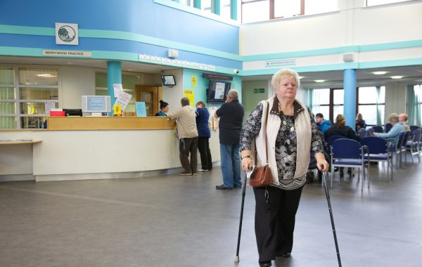 An older woman walking with sticks in a GP waiting area