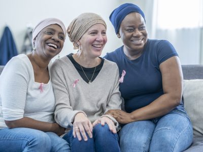 A group of three women wearing pink breast cancer awareness ribbons are sitting on a sofa, smiling