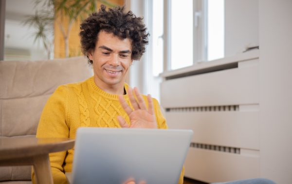A man wearing a yellow jumper, sitting on the floor of his flat with a laptop on his lap is waving to people he is having an online meeting with
