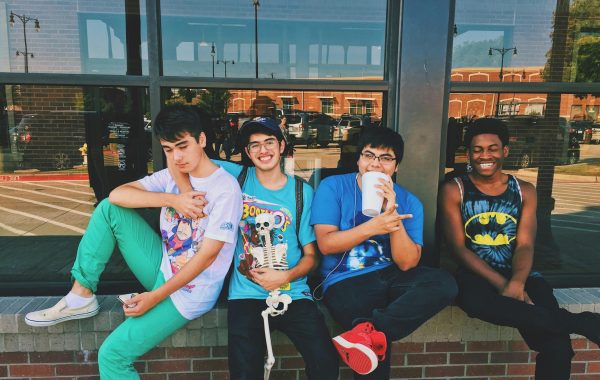Four ethnically diverse young men are sitting on a low wall with their backs against a restaurant window