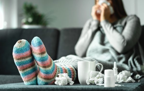 A woman sitting on a sofa with her slippered feet up on a table covered in tissues and cold remedies