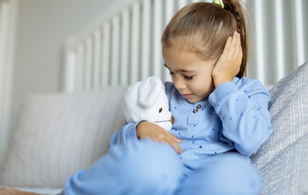 Girl on a sofa hugging a teddy and clutching her ear